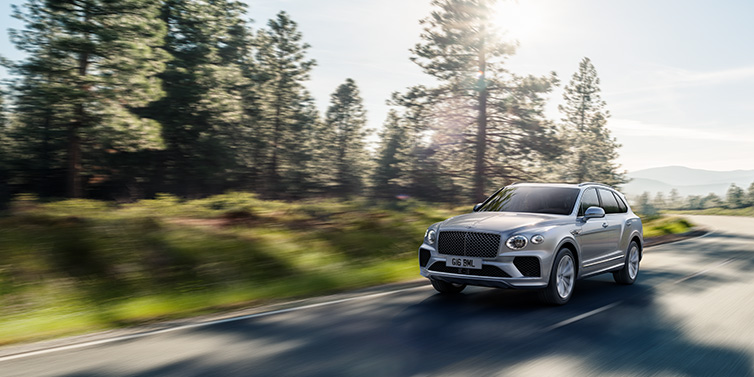 Bentley Bangkok Bentley Bentayga SUV front three quarter in Moonbeam silver paint driving dynamically along a tree-lined road