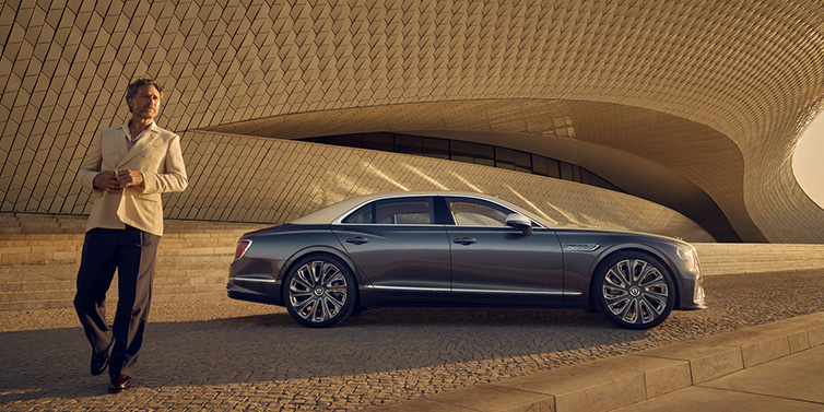 Bentley Bangkok Bentley Flying Spur Mulliner sedan in Rose Gold over Magnetic duotone paint parked by a curved wall with person in suit jacket standing on the left